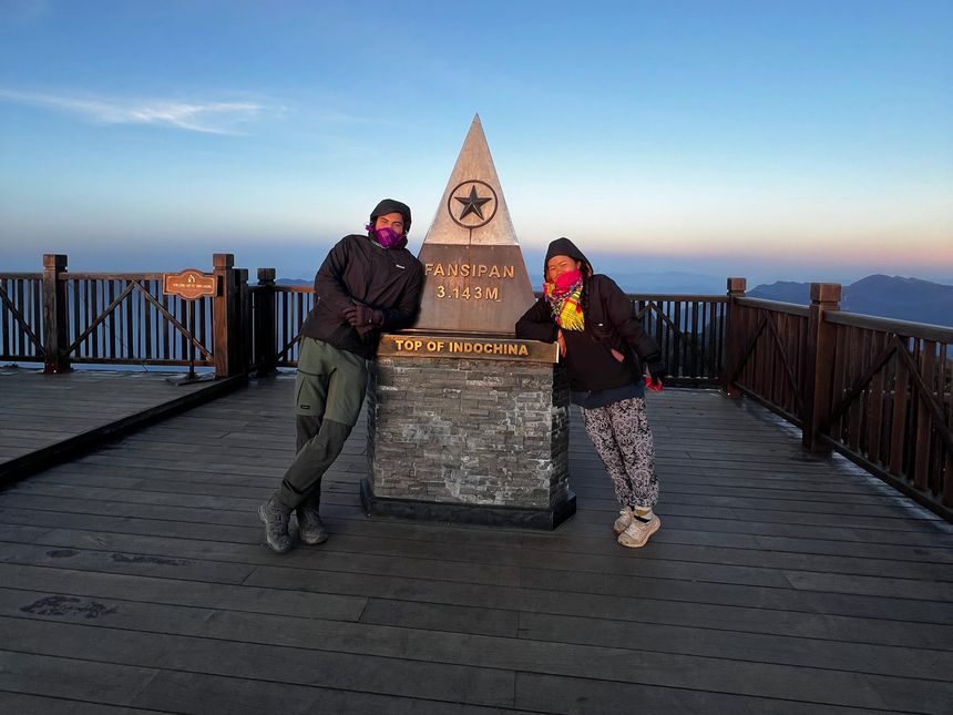 Two trekkers in warm clothes at the Fansipan summit marker (3,143 m) at dawn