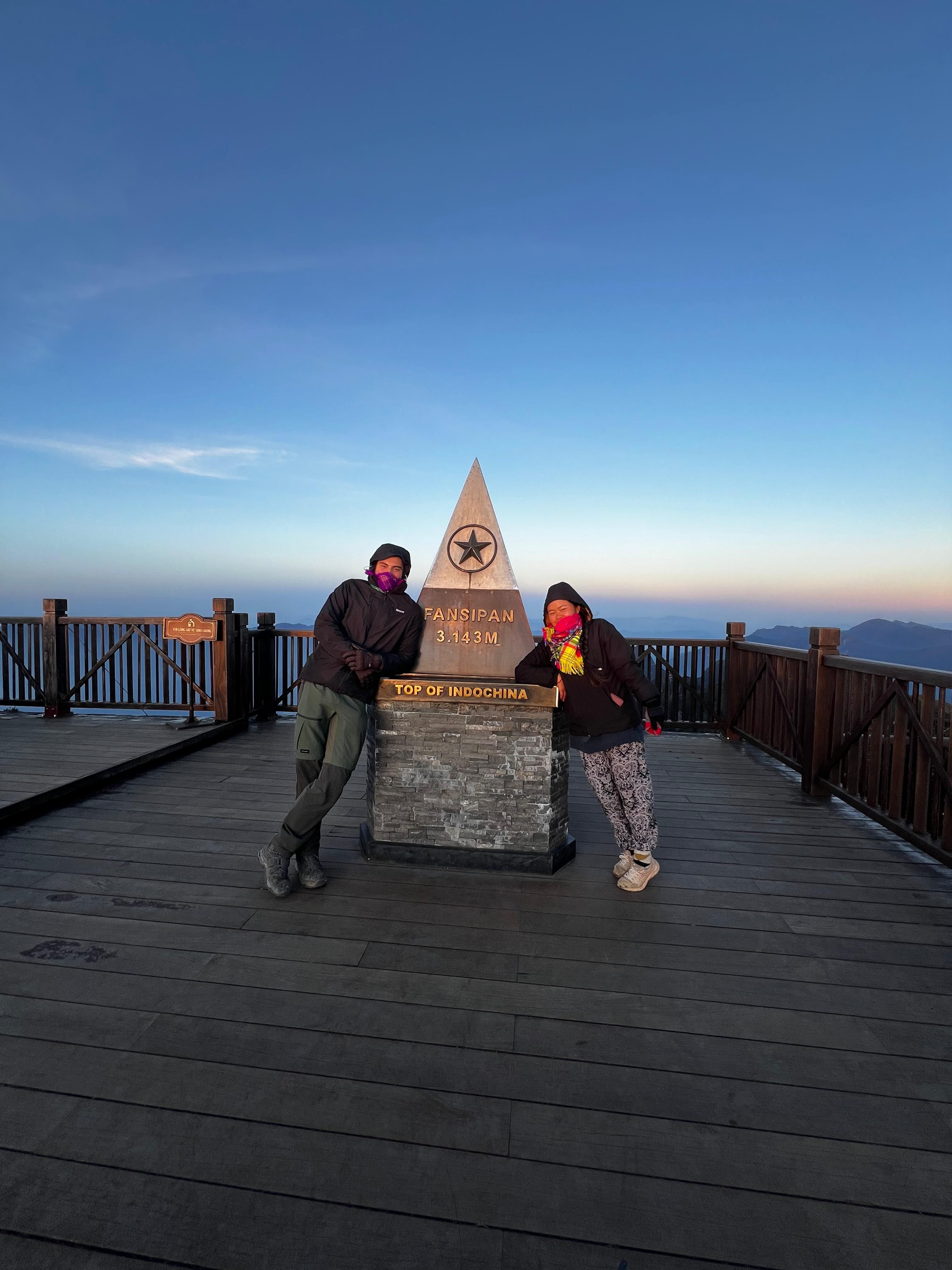 Two trekkers in warm clothes at the Fansipan summit marker (3,143 m) at dawn